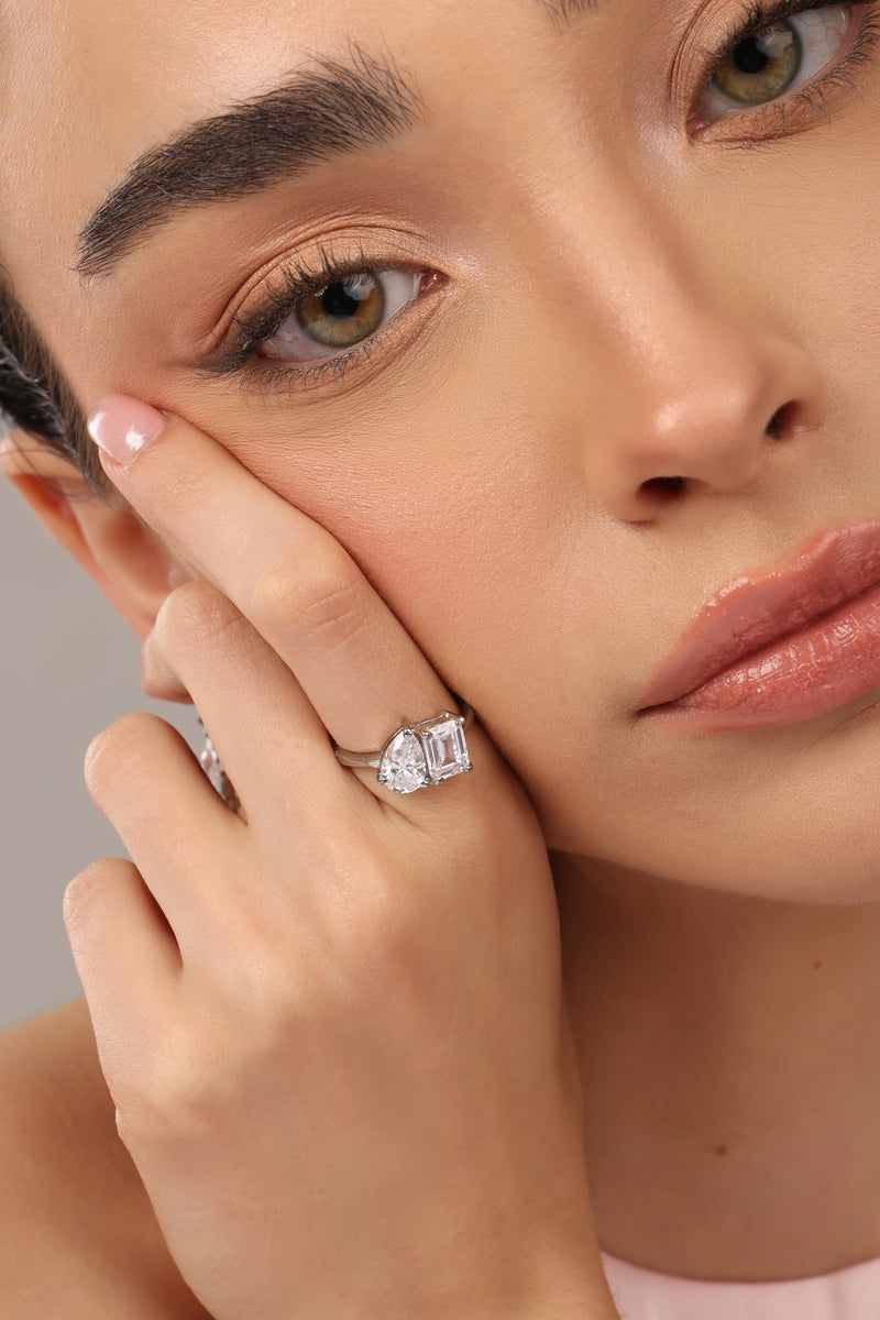 A close-up lifestyle shot of a model wearing a silver ring with three rectangular baguette-cut clear gemstones on her finger, set against a soft neutral background.